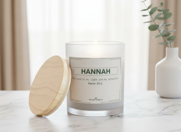 personalized frosted glass candle with a wooden lid, labeled “HANNAH” and “The Lord is my light and my salvation – Psalm 27:1” in soft green font, placed on a marble countertop with a blurred background of beige curtains and a vase with greenery, in a gentle, neutral lifestyle setting – Olive Faith Christian gift.